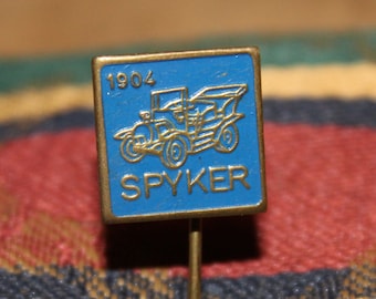 May include: A blue enamel pin with a gold border and a gold image of a vintage car. The pin says "1904" and "SPYKER" in gold lettering.