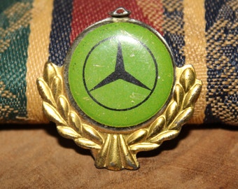 May include: A gold-colored metal pin with a green circle featuring a three-pointed star. The circle is surrounded by a wreath of gold leaves.