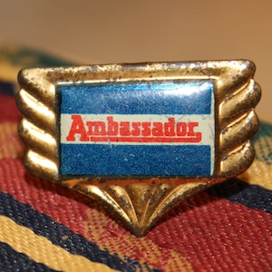 May include: A gold-colored metal pin with a blue and white enamel design. The pin features the word "Ambassador" in red letters.