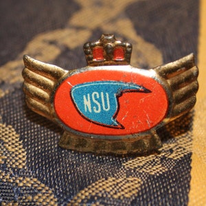 May include: A gold-coloured pin with a red and blue logo that says "NSU" and wings on either side. The pin has a crown on top.
