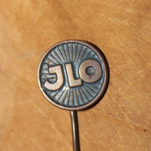 May include: A vintage JLO pin with a blue and gold design. The pin features the letters JLO in gold against a blue background with a sunburst design.