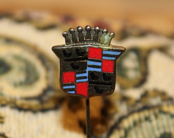 May include: A vintage gold-toned metal pin with a blue and red enamel design. The pin features a shield with a crown on top and a red and blue checkered pattern.