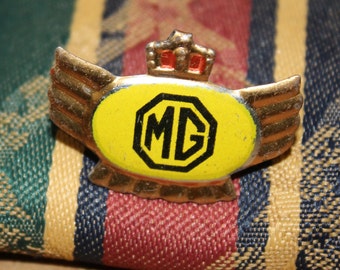 May include: A gold-colored metal pin with a yellow oval background and a black and white MG logo. The pin has a crown on top and wings on the sides.