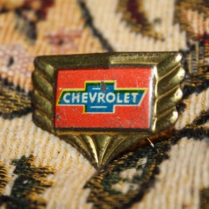 May include: A vintage Chevrolet emblem pin with a gold-coloured frame and a red background. The pin features the Chevrolet logo in white and blue. The pin is a small, rectangular shape.
