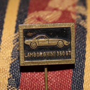 Könnte beinhalten: Eine goldfarbene Anstecknadel mit schwarzem Hintergrund, auf dem ein weißer Umriss eines Lamborghini 350GT-Autos zu sehen ist. Der Text "Lamborghini 350GT" ist in weißen Buchstaben unter dem Auto geschrieben.