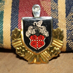 May include: A vintage metal charm featuring a black and red shield with a white bird and a crown. The shield is surrounded by a gold-colored wreath.