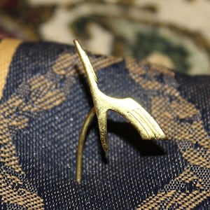 May include: A gold-colored metal pin shaped like a stylized bird with outstretched wings.