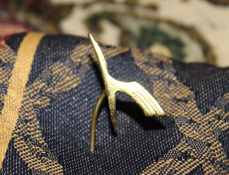 May include: A gold-colored metal pin shaped like a stylized bird with outstretched wings.