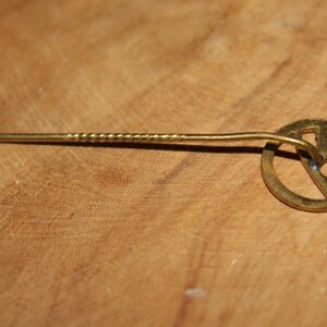 May include: A gold-colored pin with a circular design featuring a stylized letter 'R' inside. The pin has a twisted metal stem.