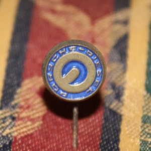May include: A vintage pin with a circular design. The pin features a blue and gold color scheme with the word "DIESEL" around the edge. The center of the pin has a blue "S" shape. The pin is set against a patterned fabric background.