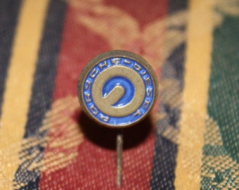 May include: A vintage pin with a circular design. The pin features a blue and gold color scheme with the word "DIESEL" around the edge. The center of the pin has a blue "S" shape. The pin is set against a patterned fabric background.