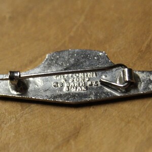 May include: Silver-toned metal pin with a design of a ship. The back of the pin has the text "TATLOCKINI & SONS 55 PARK ST. SWAN"