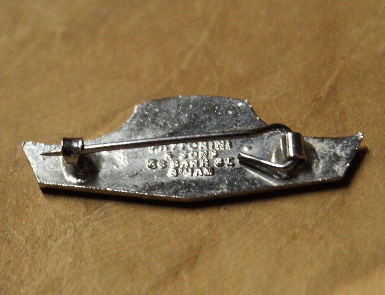 May include: Silver-toned metal pin with a design of a ship. The back of the pin has the text "TATLOCKINI & SONS 55 PARK ST. SWAN"