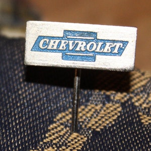 May include: A silver pin with a blue and white Chevrolet logo. The pin is on a brown and black fabric.