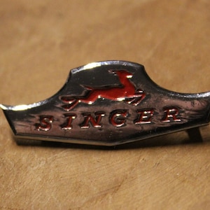 May include: A silver and red Singer sewing machine badge with a stylized running deer logo and the word "SINGER" in raised letters.