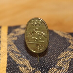 May include: A gold-colored pin with the Olympic rings and a depiction of a horse rider. The pin says "Tokyo 2004" on it.