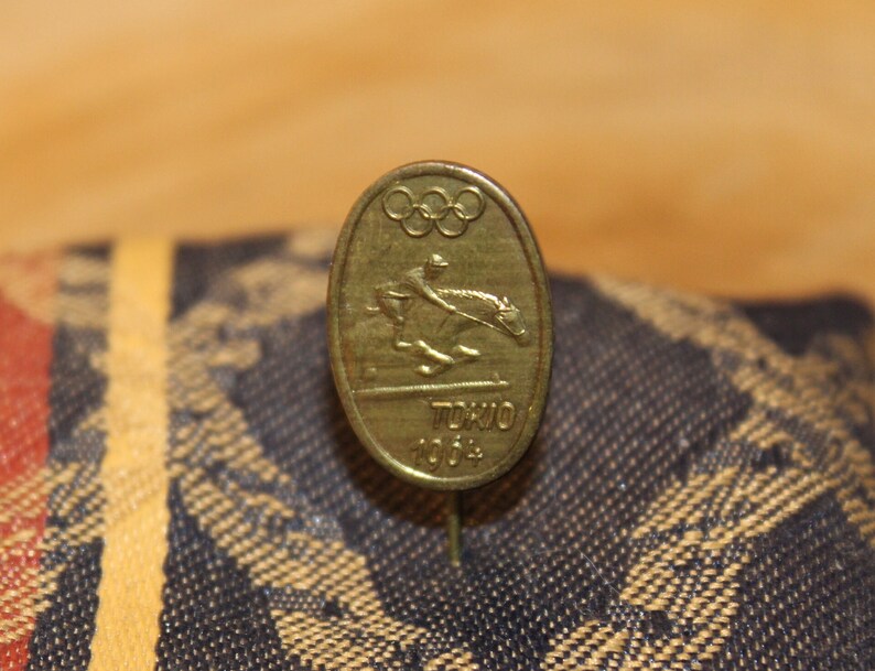 May include: A gold-colored pin with the Olympic rings and a depiction of a horse rider. The pin says "Tokyo 2004" on it.