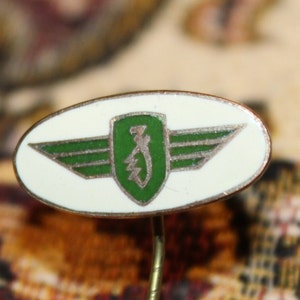 May include: Oval-shaped pin with a white background and a green emblem featuring wings and a shield. The pin has a gold-colored metal border and a pin backing.