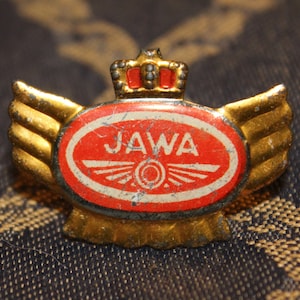 May include: A gold-colored metal pin with a red and white oval emblem featuring the word "JAWA" and a stylized winged wheel. The pin has a crown on top and wings on either side.