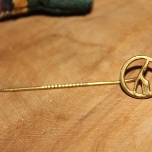 May include: A gold-toned pin with a peace sign design featuring a stylized giraffe inside. The pin has a long, thin, twisted metal stem.