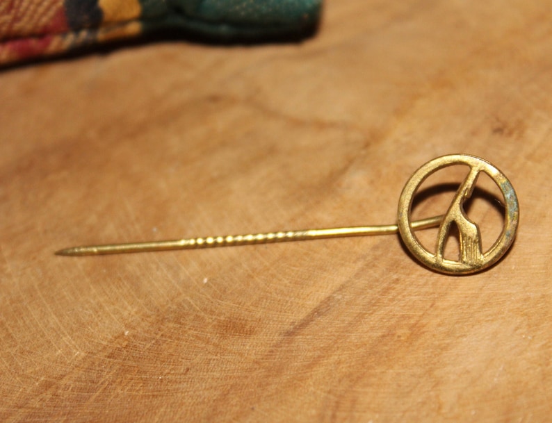 May include: A gold-toned pin with a peace sign design featuring a stylized giraffe inside. The pin has a long, thin, twisted metal stem.