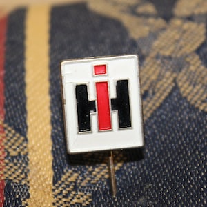 May include: A white enamel pin with a red and black IH logo. The pin is on a fabric background.