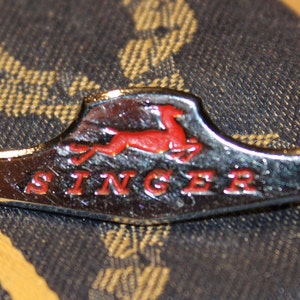 May include: A silver and red Singer sewing machine badge with a stylized red leaping deer logo and the word "SINGER" in red lettering.