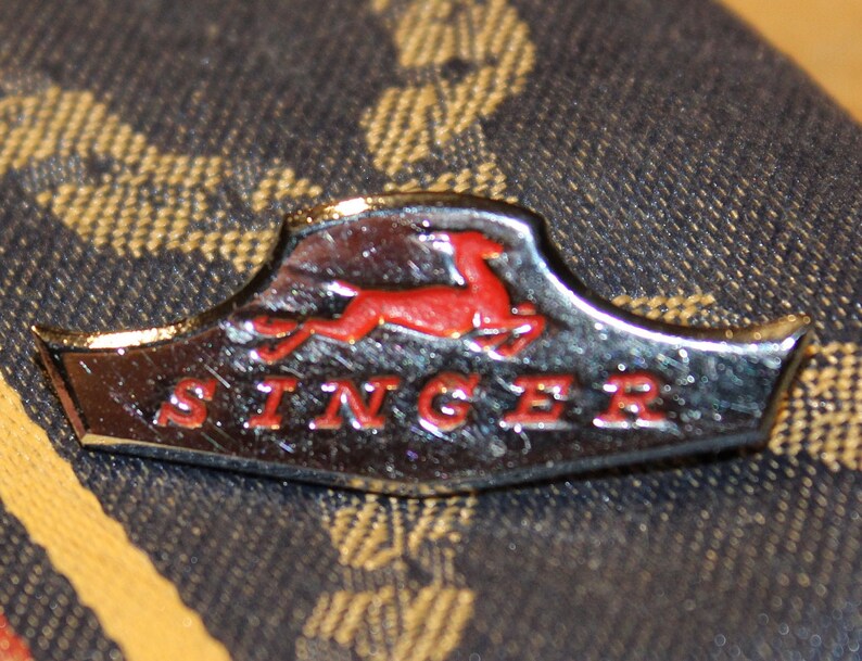 May include: A silver and red Singer sewing machine badge with a stylized red leaping deer logo and the word "SINGER" in red lettering.