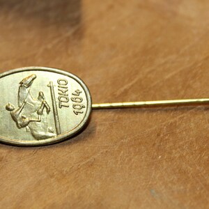 May include: Gold-colored pin with an oval-shaped design featuring the Olympic rings and a rider on horseback. The text "TOKYO 1964" is inscribed on the pin.