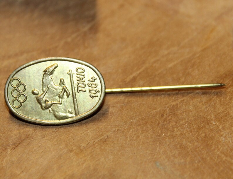 May include: Gold-colored pin with an oval-shaped design featuring the Olympic rings and a rider on horseback. The text "TOKYO 1964" is inscribed on the pin.