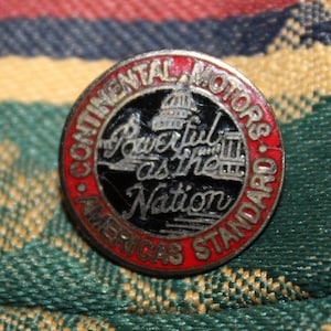 May include: A red, white, and black enamel pin with the text "Continental Motors - America's Standard" and the words "Powerful as the Nation" in a silver script font. The pin features a black and white image of the Capitol Building.