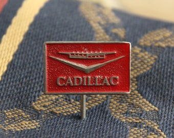 May include: A red enamel pin with a silver Cadillac logo and the word "CADILLAC" in silver lettering.