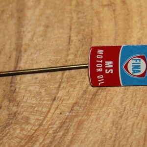 May include: A vintage FINA MS Motor Oil lapel pin. The pin features a rectangular design with a red and blue color scheme, and the text "MS MOTOR OIL" is printed vertically. The pin has a gold-colored metal pin.