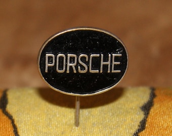 May include: A vintage Porsche pin with a black oval center and the brand name in silver lettering. The pin has a silver border and a silver pin backing. The background is a blurred mix of colors.