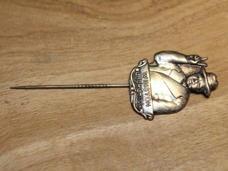 May include: Silver lapel pin featuring a sculpted image of Winston Churchill with the text "Winston Churchill" on the side. The pin has a long, thin, silver pin back.