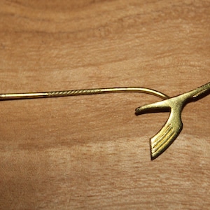 May include: A gold-colored pin with a stylized bird design. The pin has a long, thin stem with a pointed end.