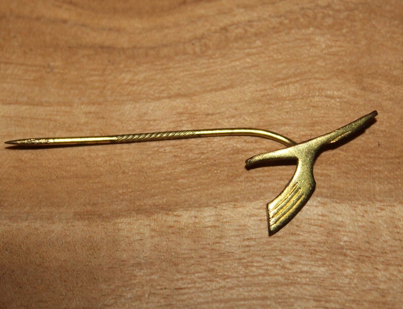 May include: A gold-colored pin with a stylized bird design. The pin has a long, thin stem with a pointed end.