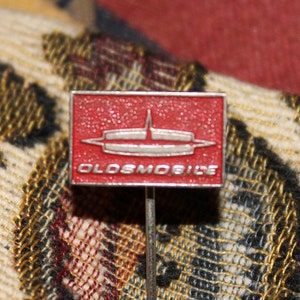 May include: A red and silver pin with the word "OLDSMOBILE" in white letters. The pin features a stylized rocket ship design.