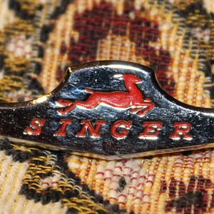 May include: A silver and red Singer sewing machine logo with a stylized running deer and the word "SINGER" in red letters.