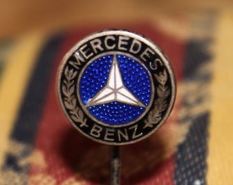May include: A silver pin with a blue and white Mercedes-Benz logo. The logo is a three-pointed star inside a circle with the words "Mercedes-Benz" around it.