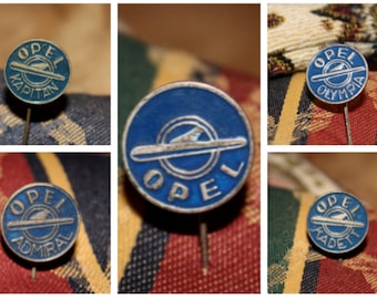May include: Four vintage blue and silver Opel car model pins. The pins feature the Opel logo and the model names: Kapitän, Olympia, Admiral, and Kadett.