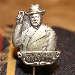 May include: Silver-toned lapel pin featuring a portrait of Winston Churchill in a hat, giving a peace sign. The pin has a banner with the text "Winston Churchill".