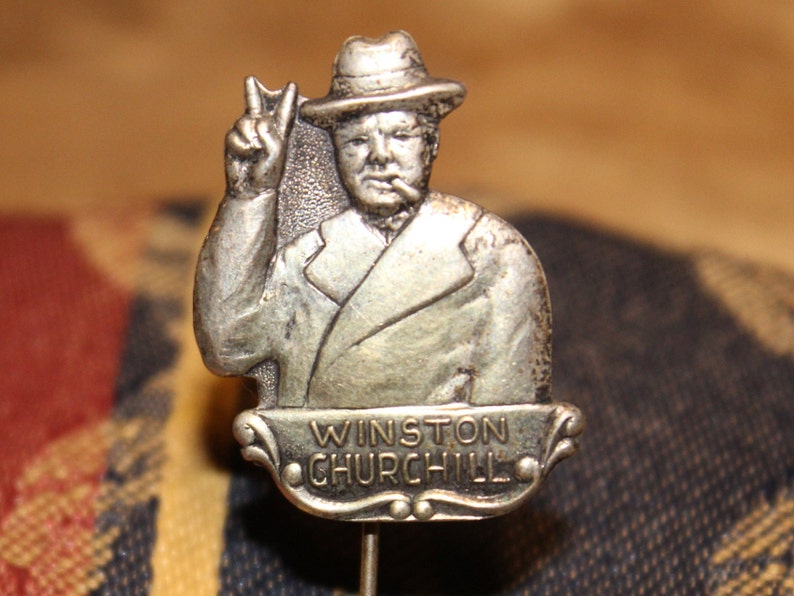 May include: Silver-toned lapel pin featuring a portrait of Winston Churchill in a hat, giving a peace sign. The pin has a banner with the text "Winston Churchill".