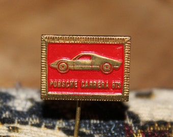 May include: A rectangular gold-tone metal lapel pin featuring a red enamel background. The pin displays a raised gold-colored silhouette of a sports car and the embossed text PORSCHE CARRERA GTS. It has a textured border and a thin metal pin backing.