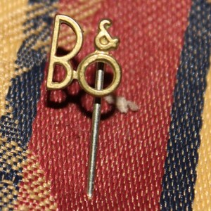 Può includere: Una spilla da bavero in metallo color oro con le lettere stilizzate B e O collegate da una e commerciale. Il design include un lungo spillo metallico dritto e sottile di colore argento che si estende verso il basso dalla base della lettera O.