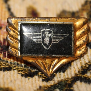 May include: A gold-colored metal pin with a black enamel background and a silver-colored eagle with wings spread. The eagle is inside a shield shape.