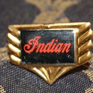 May include: Gold-colored metal pin with a black background and the word "Indian" in red lettering. The pin has a wing-like design.