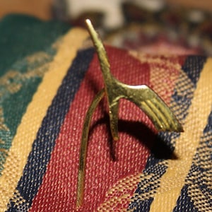May include: A gold-colored metal bird figurine with a long, curved neck and a detailed wing design. The figurine is sitting on a striped fabric surface.