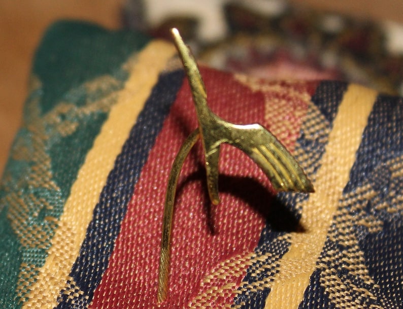 May include: A gold-colored metal bird figurine with a long, curved neck and a detailed wing design. The figurine is sitting on a striped fabric surface.