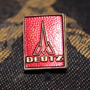 Może przedstawiać: Metalowa szpilka w kolorze czerwonym i złotym z napisem "DEUTZ" i stylizowanym wzorem pasma górskiego.